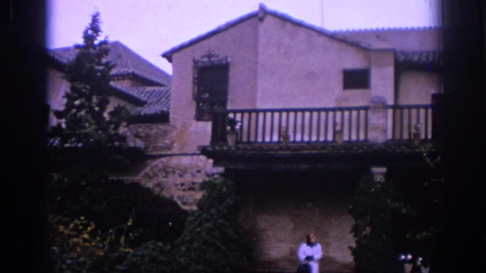 1966 Spain 8mm frame showing the balcony-fronted building, evergreen trees, courtyard planting, and a woman in light clothing below.