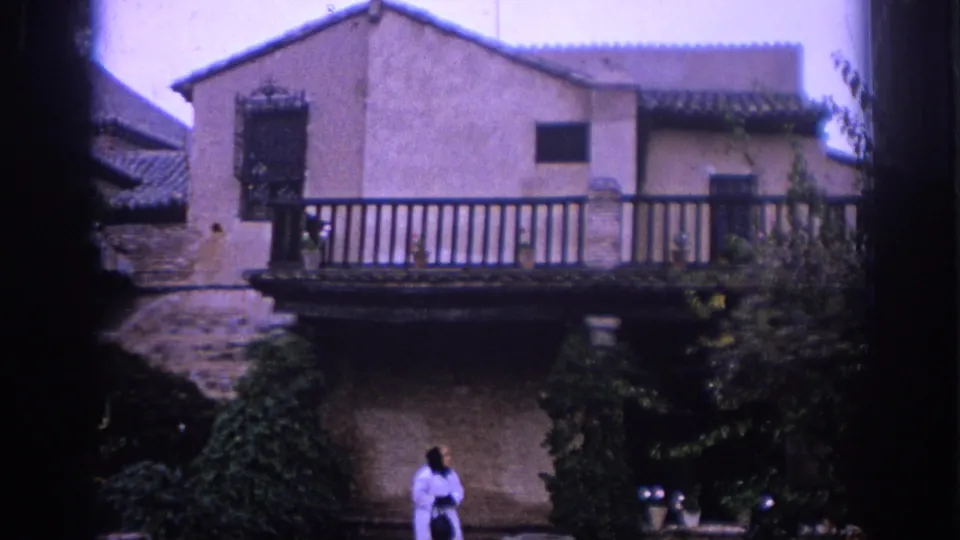 1966 Spain home-movie frame with a woman below a balcony, surrounded by courtyard greenery and a stone-and-stucco building facade.