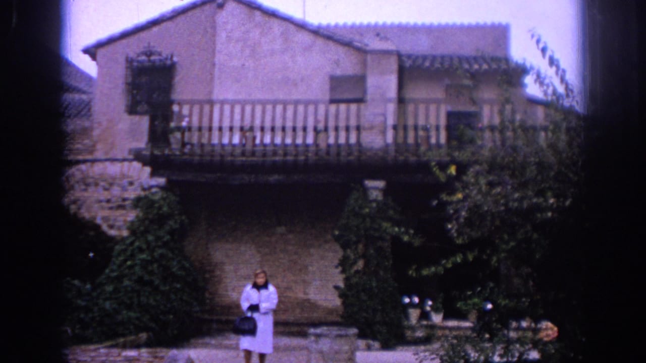 Adult woman standing before a stone building with a wooden balcony and greenery.