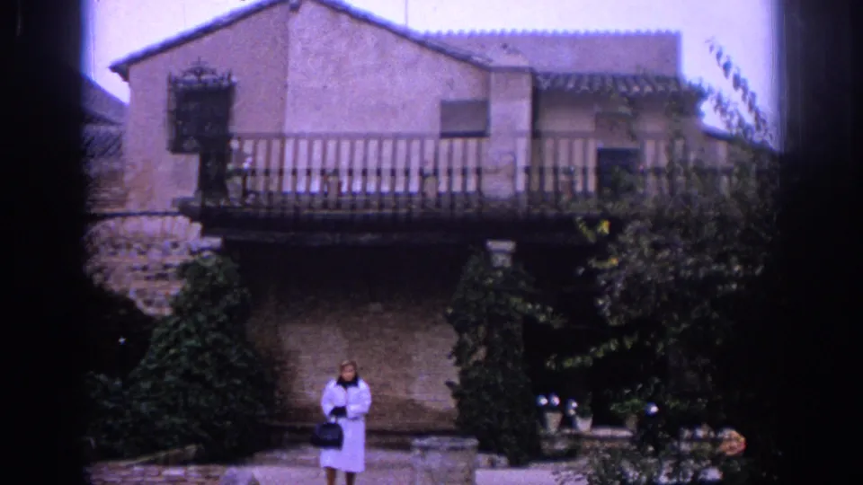 1966 Spain home-movie frame showing an adult woman near a stone building with a wooden balcony, plants, and shaded facade.