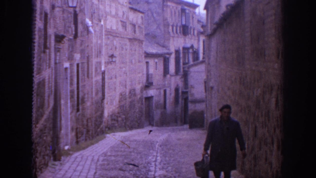 1966 Spain street view with an adult man carrying a bag along a narrow, sloped lane lined by weathered stone facades.