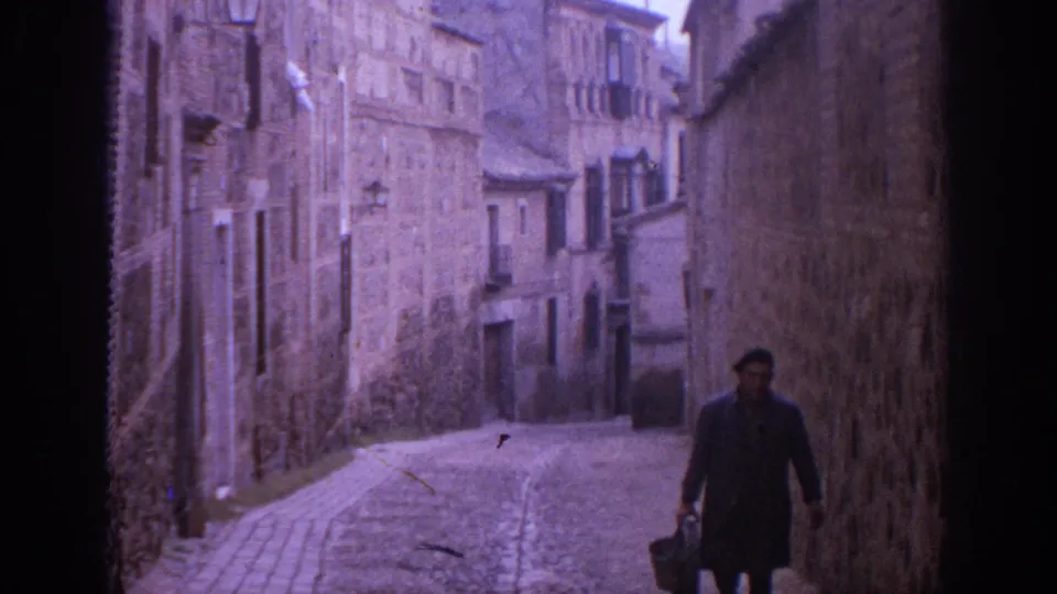 1966 Spain street view with an adult man carrying a bag along a narrow, sloped lane lined by weathered stone facades.