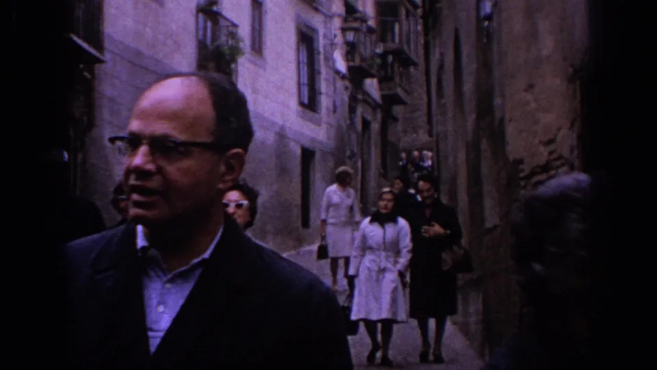 1966 Spain home-movie frame with adult tourists in coats walking through a narrow historic street behind a man in glasses.