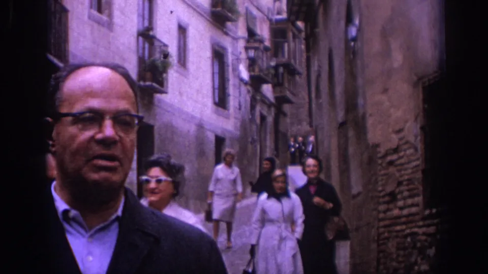 1966 Spain travel frame showing a man in glasses in the foreground while several adult tourists walk behind him through a tight stone-lined street.
