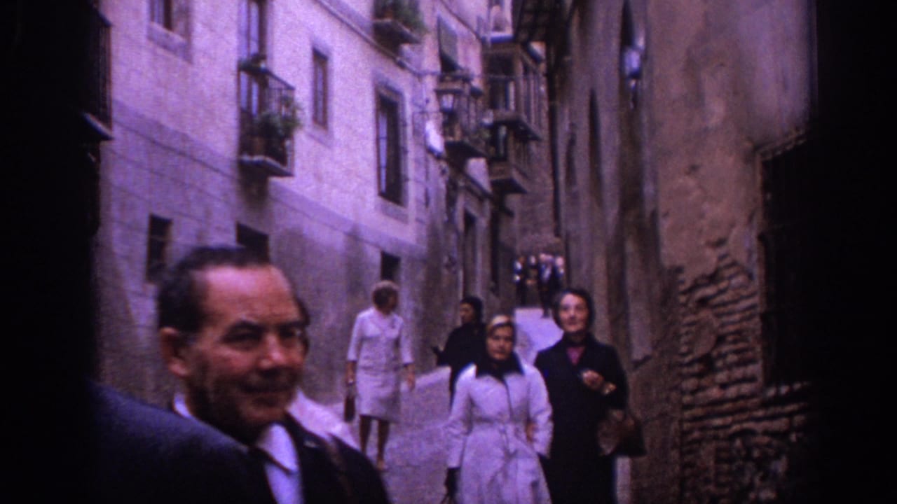 In 1966 Spain, several pedestrians in period clothing walk through a narrow stone alley lined with old buildings.