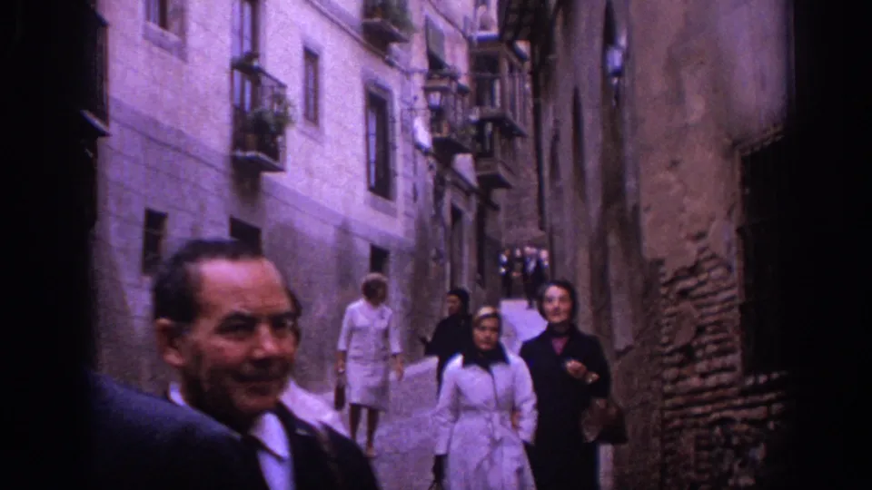 In 1966 Spain, several pedestrians in period clothing walk through a narrow stone alley lined with old buildings.