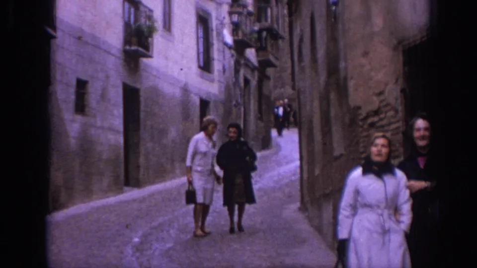 Two women in mid-1960s clothing walk down a narrow Spanish alley with other pedestrians visible near the buildings.