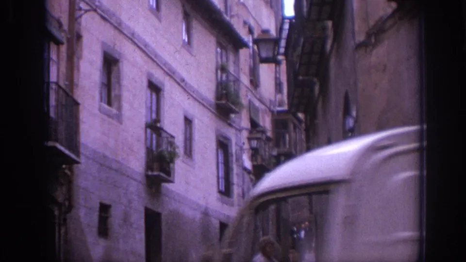 The edge of a vehicle passes in the foreground beside weathered buildings and balconies in the Spanish street scene.