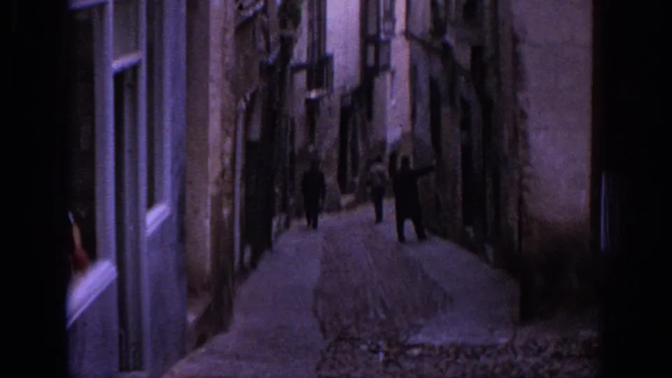 1966 Spain home-movie view down a narrow cobblestone alley lined with old buildings and small figures walking away.