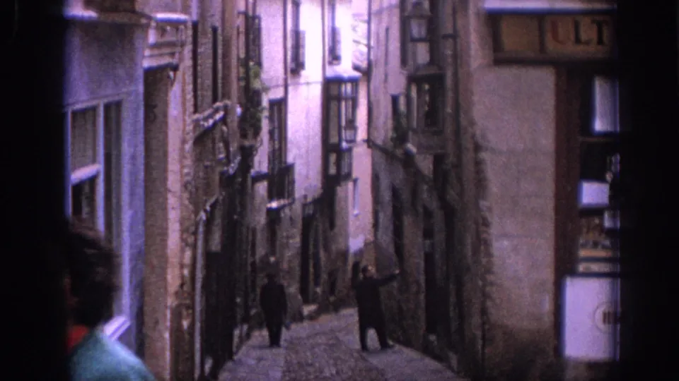 A foreground figure in green passes near the camera as the 1966 Spain footage looks down a narrow historic street.