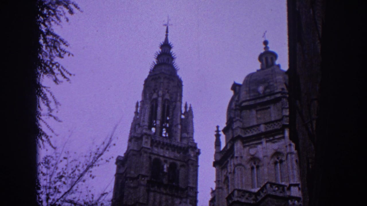 1966 Spain home-movie frame showing Gothic cathedral towers and stone facade seen upward through dark trees under a purple-blue sky.