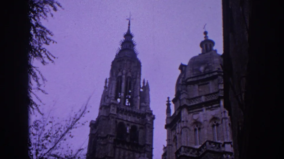 1966 Spain home-movie frame showing Gothic cathedral towers and stone facade seen upward through dark trees under a purple-blue sky.