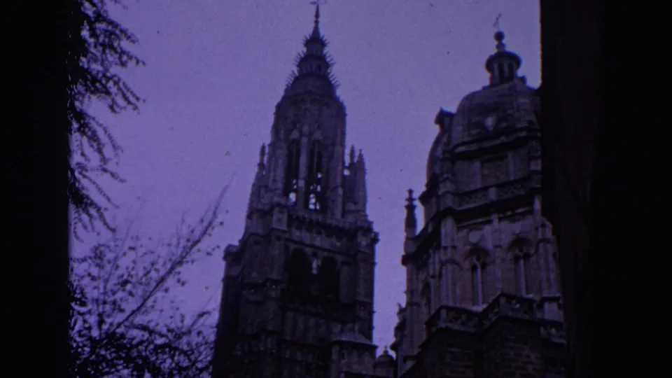 1966 Spain frame with a tall Gothic tower, adjacent domed roof feature, bare branches, and dark foreground tree trunks.