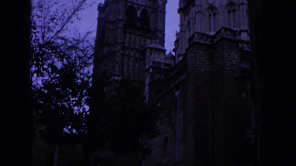 1966 Spain 8mm frame showing a shadowed Gothic cathedral tower and stone wall through trees, with a cool purple cast.