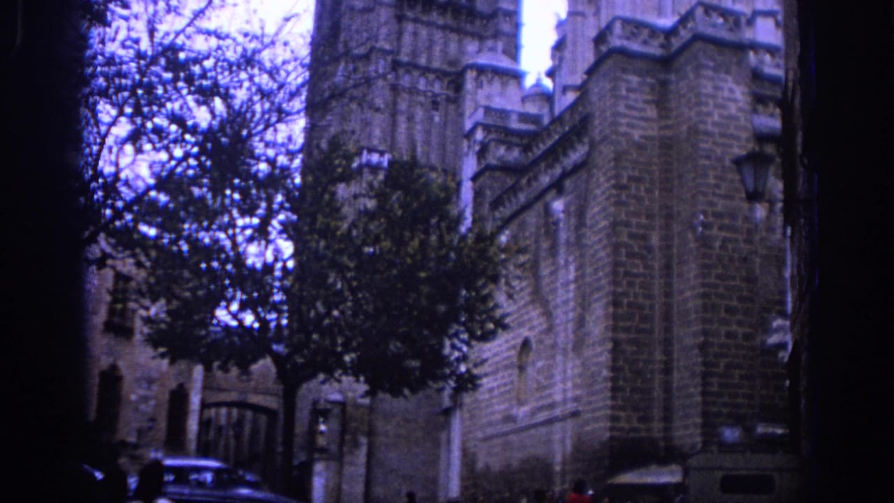 1966 Spain 8mm frame showing a Gothic cathedral exterior seen from a nearby street with trees, masonry, and parked cars.