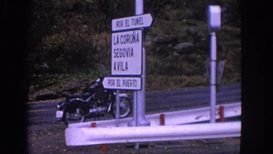 1966 Spain 8mm roadside view with signs for La Coruna, Segovia, and Avila beside a parked motorcycle.