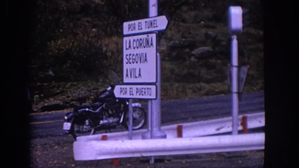 1966 Spain roadside frame showing a white signpost for La Coruna, Segovia, and Avila with a motorcycle nearby.
