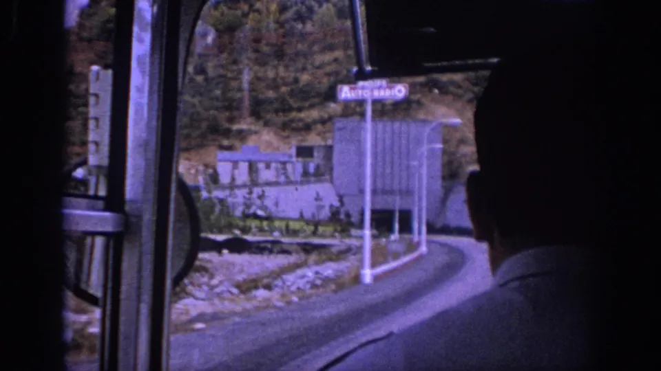 Interior bus perspective in Spain showing window frames, a passenger silhouette, a road bend, and a building near the tunnel approach.