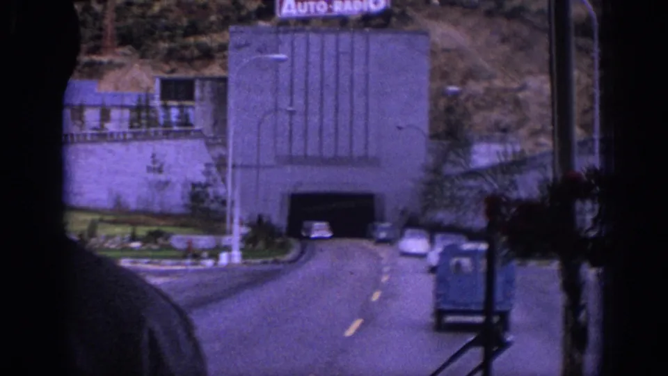 1966 Spain home-movie view from inside a vehicle approaching a tunnel, with mid-century cars, a small blue truck, and hillside buildings visible.