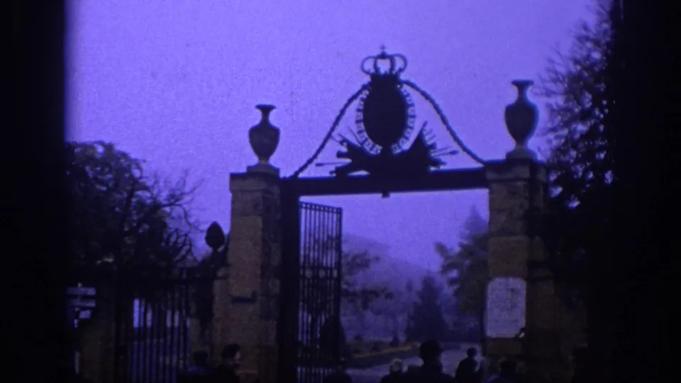 1966 Spain home-movie frame of an ornate iron gate with stone posts, crown emblem, trees, and small visitors gathered near the entrance.