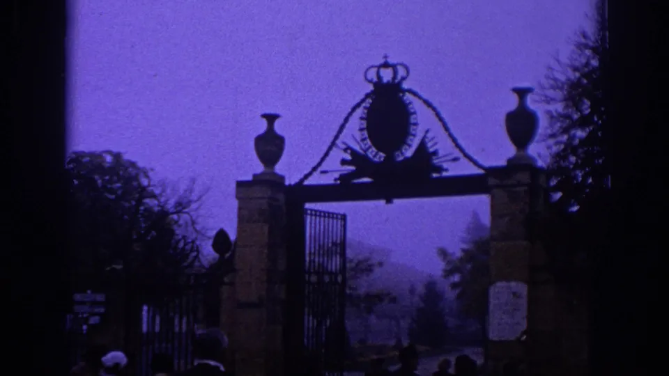 1966 Spain frame showing a formal stone-and-iron gate with crown ornament, trees, and visitors partly visible near the bottom of the shot.