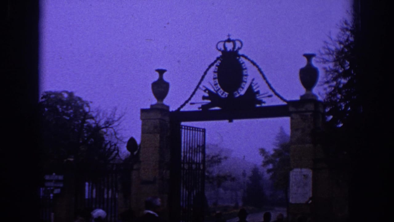 1966 Spain 8mm frame showing a formal park entrance with stone posts, urns, iron fencing, and a crowned crest in purple haze.