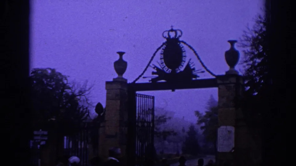 1966 Spain 8mm frame showing a formal park entrance with stone posts, urns, iron fencing, and a crowned crest in purple haze.