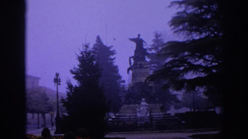1966 Spain home-movie frame showing a hazy park scene with evergreens, iron fencing, and a mounted statue beyond the path.