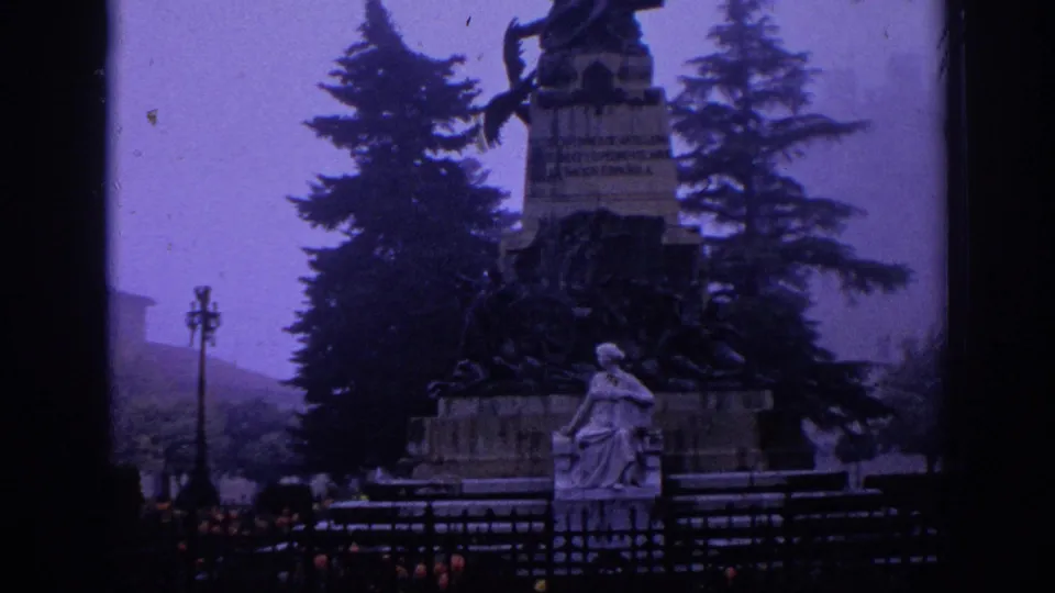 1966 Spain 8mm frame revealing more of the monument base, seated sculpture, railing, trees, and hazy hillside beyond.