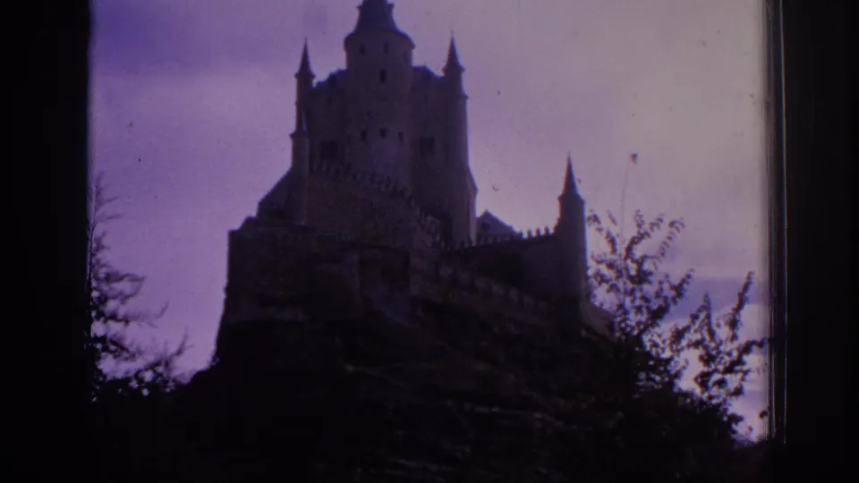 1966 Spain 8mm frame holding on the castle silhouette, with turrets, battlements, and dark foliage beneath a violet sky.