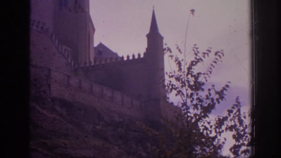 A Spanish castle exterior appears through 8mm film framing, with battlements, a pointed turret, hillside vegetation, and muted color shift.
