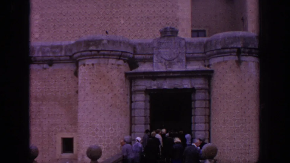 1966 Spain travel footage frame showing visitors gathered at a stone castle doorway below patterned brickwork and rounded facade details.