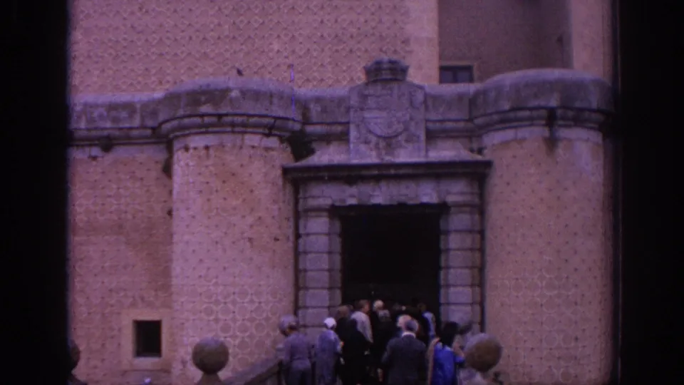 Visitors in mid-1960s attire stand at the dark entrance of a Spanish castle, with brick-patterned walls and stone trim visible.
