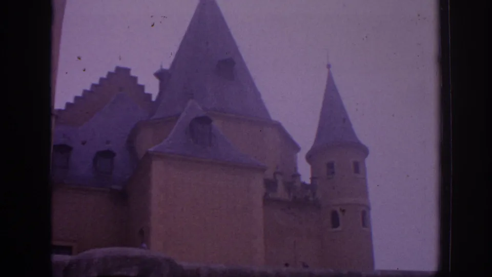 1966 Spain home-movie frame showing castle turrets, steep conical roofs, and soft purple-tinted sky.