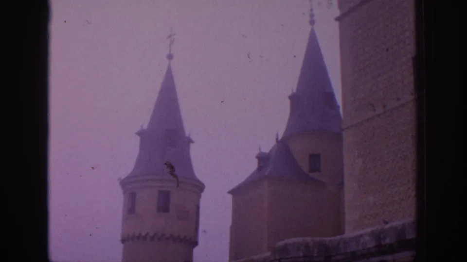 Purple-cast 8mm view of Spanish castle towers with pointed roofs and a close stone wall at right.