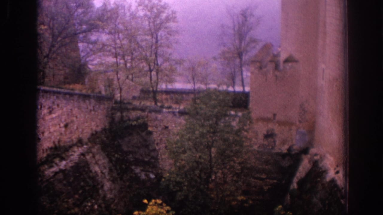 1966 Spain 8mm frame looking from a shaded interior or balcony toward stone courtyard walls, trees, and part of a tower.