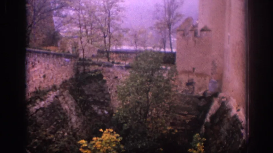 1966 Spain travel footage frame showing a stone wall, tower face, trees, shrubs, and shadowed foreground edges from a high viewpoint.