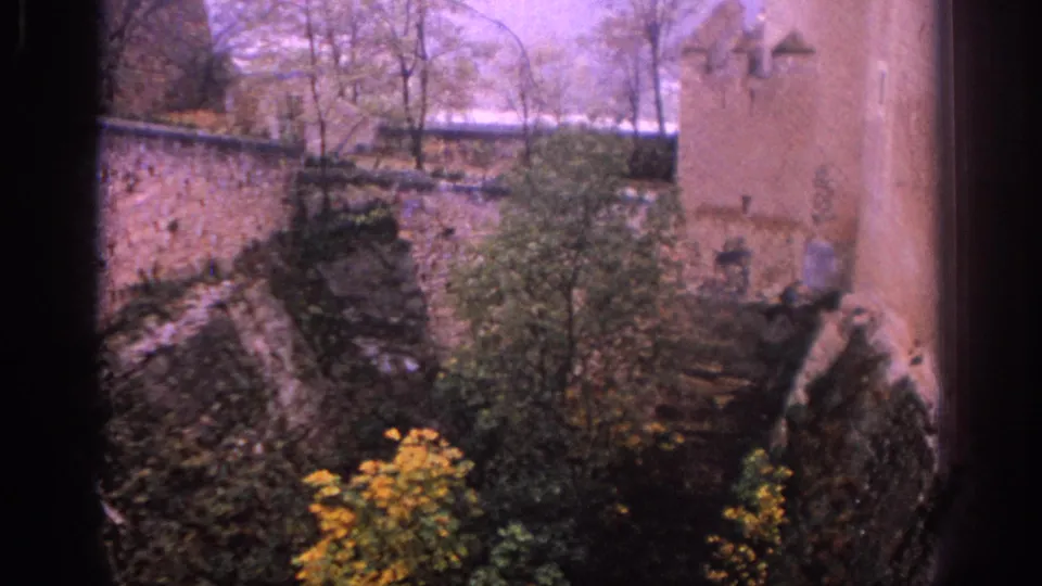 1966 Spain 8mm frame from an elevated overlook, showing old stone walls, a tower side, trees, shrubs, and yellow flowers.