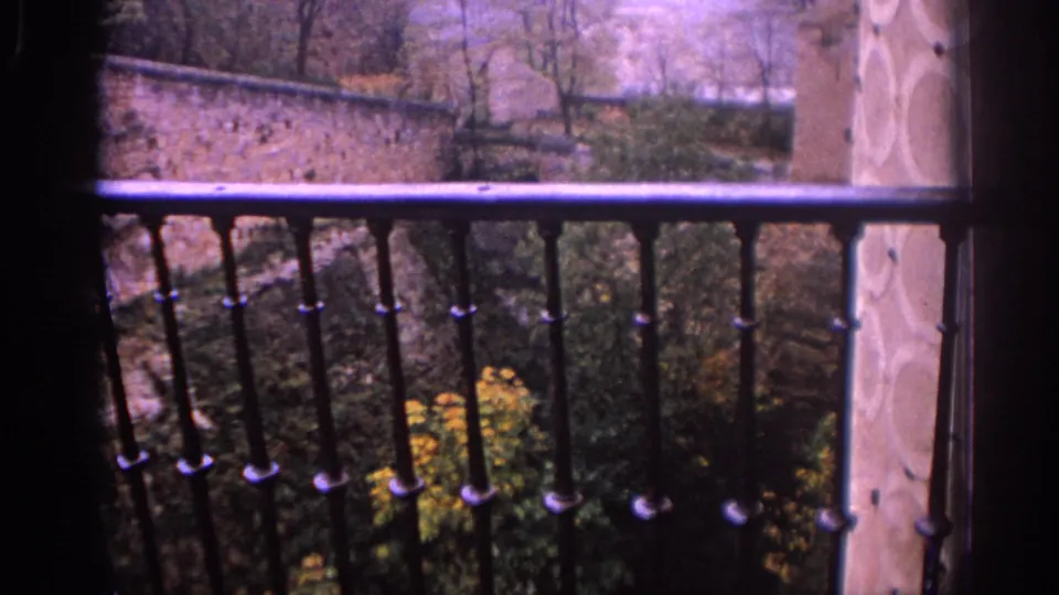 1966 Spain frame revealing a metal balcony railing in the foreground, with stone walls, trees, flowers, and a tower wall beyond.