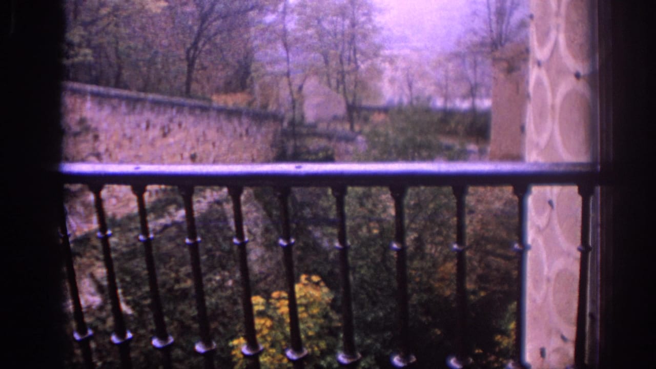 1966 Spain 8mm frame looking past an iron balcony railing toward trees, a stone wall, and muted foliage.