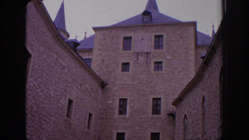 1966 Spain home-movie frame with textured stone walls, narrow windows, roof spires, and a central castle facade seen through an arch.