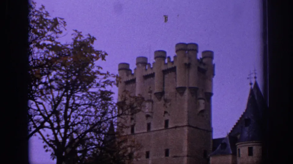 1966 Spain home-movie view of a castle tower with battlements, flanked by tree branches and nearby roof sections beneath a purple sky.