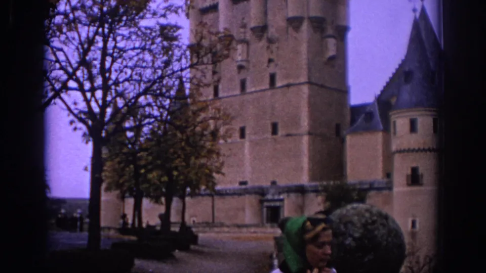 1966 Spain 8mm frame showing a woman in a green headscarf and sunglasses near a castle courtyard and trees.