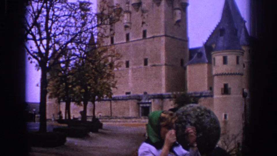 1966 Spain home-movie frame with a woman adjusting her headscarf near a tree-lined castle courtyard.