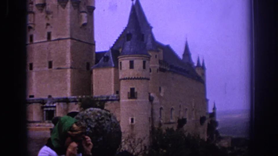 1966 Spain home-movie frame showing a woman in sunglasses and green headscarf near a large stone castle with turrets.