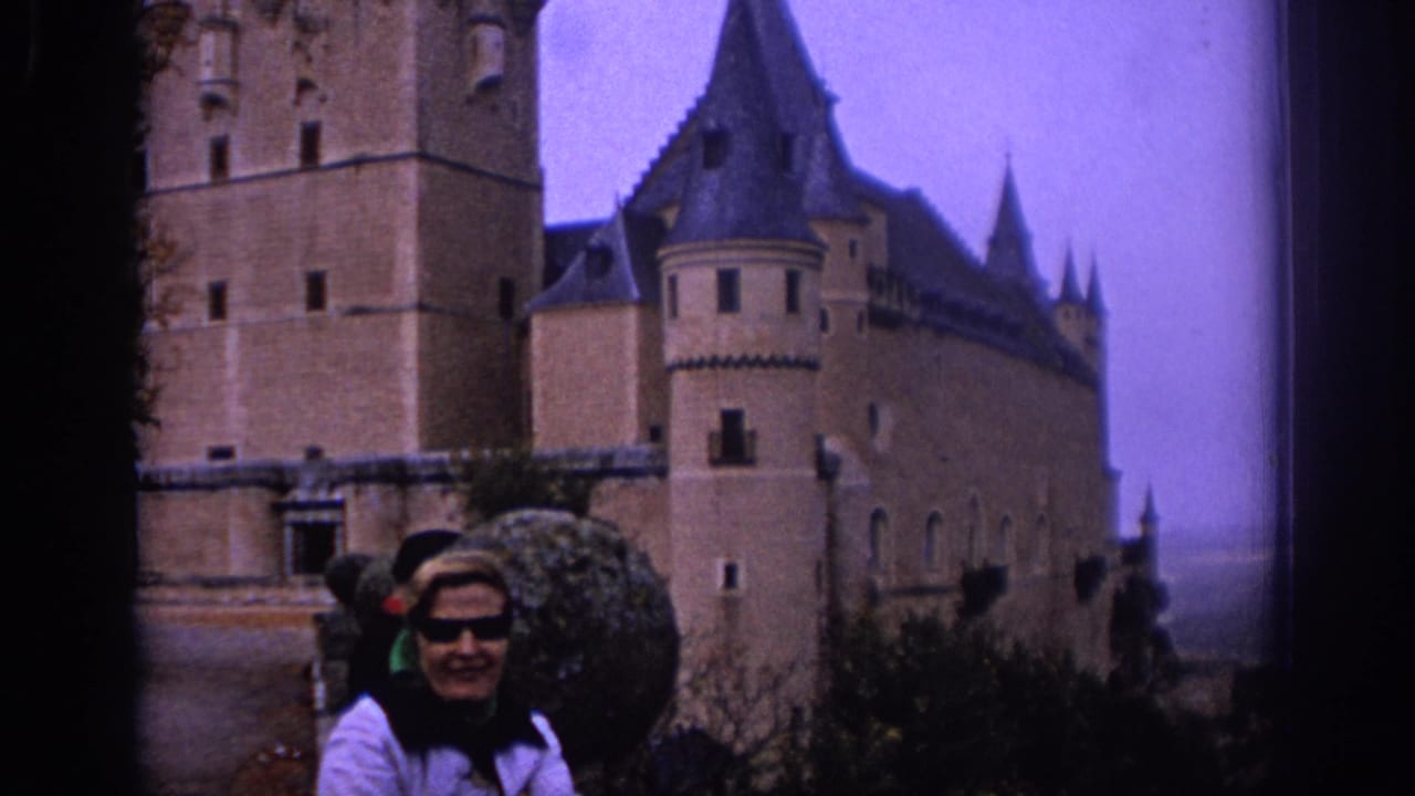 Smiling woman in sunglasses stands near a round shrub with castle walls behind her.
