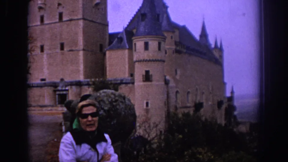 1966 Spain travel frame showing a woman facing the camera before a large stone castle with towers and steep roofs.