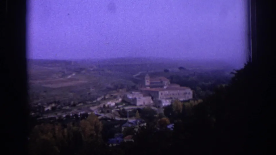 1966 Spain home-movie frame showing a church complex on a wooded rise, with fields and haze in the distance.