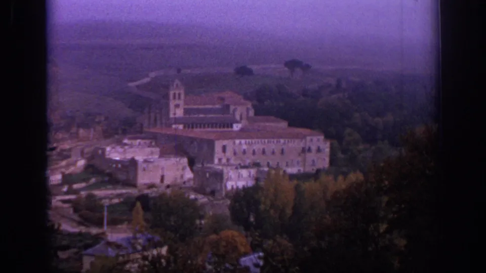 Closer 1966 Spain 8mm view of a church, tower, attached buildings, trees, and nearby stone structures.