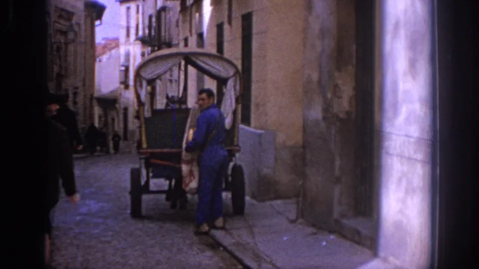 A man in blue work clothes stands with a horse-drawn carriage on a narrow cobblestone street in 1966 Spain.